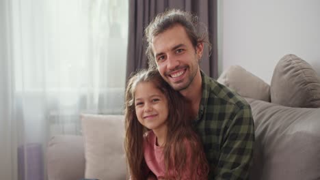 retrato de un padre feliz de un hombre moreno en una camisa a cuadros verde que está sentado con su hija pequeña, una chica morena en un vestido rosado en un sofá gris y abrazando en un apartamento moderno