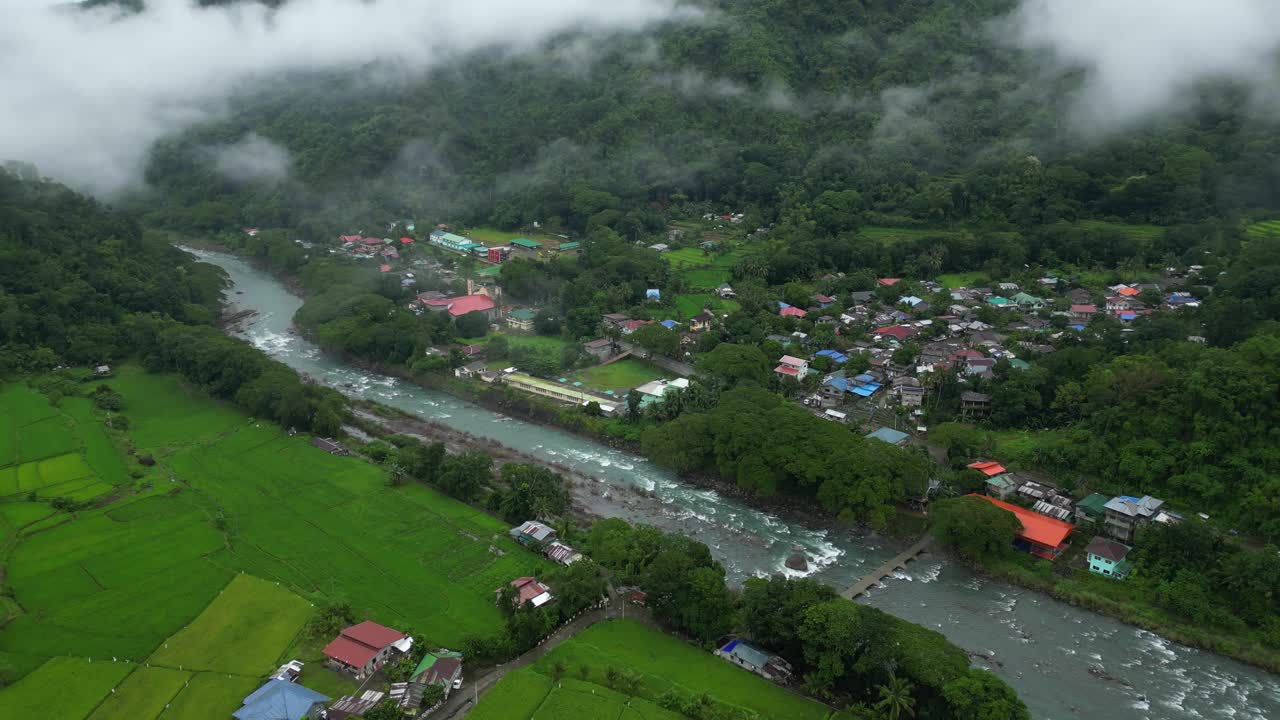 A wide aerial sweep a lush Philippine valley, revealing rice fields, a flowing river, misty forested hills, and a vibrant town with colorful rooftops