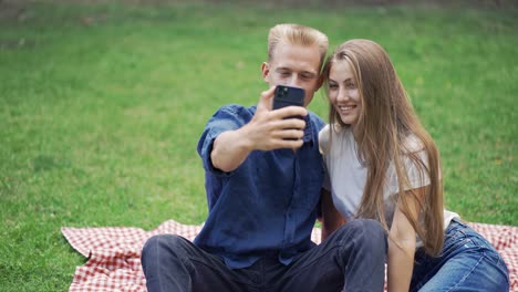 a guy and a girl on a picnic take a selfie. people sit on a mat in the park. happiness, leisure