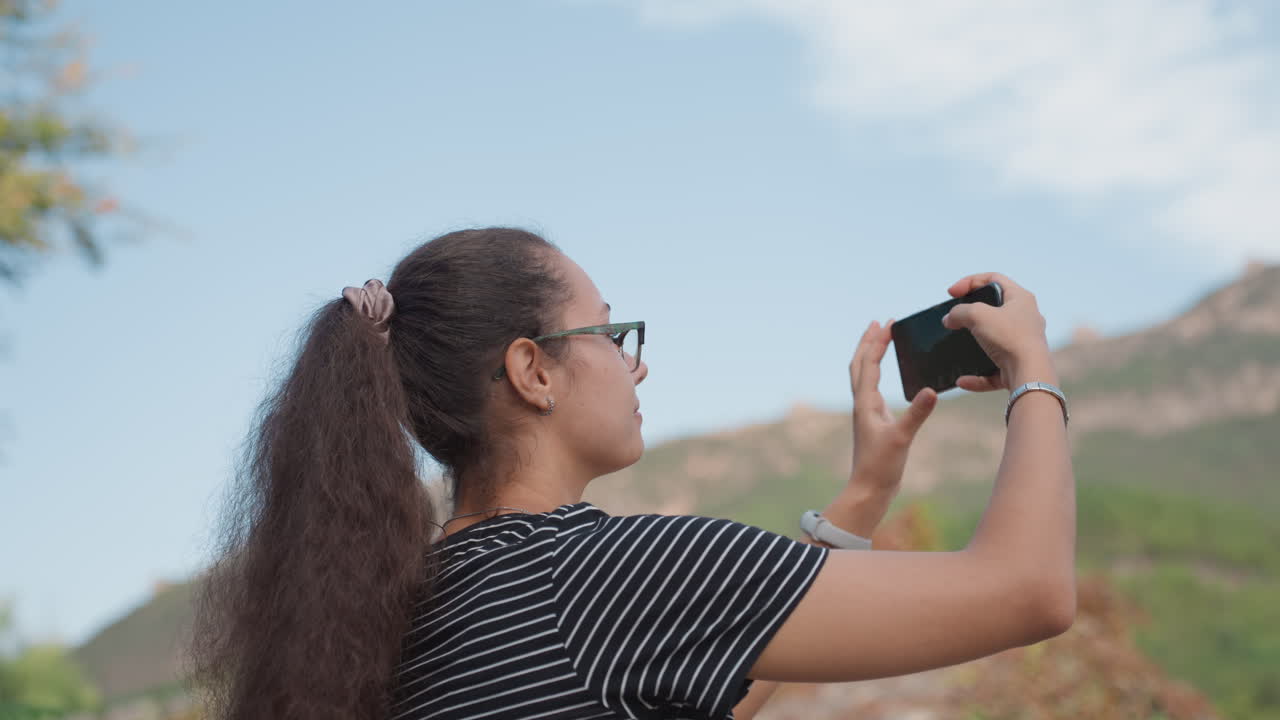 Viaggiatrice che fotografa il cielo di montagna con lo smartphone, primi piani catturano energia entusiasta, camicia a righe, occhiali, capelli ricci, dettagli di bracciale e orologio, primo piano foglioso e tetti di templi