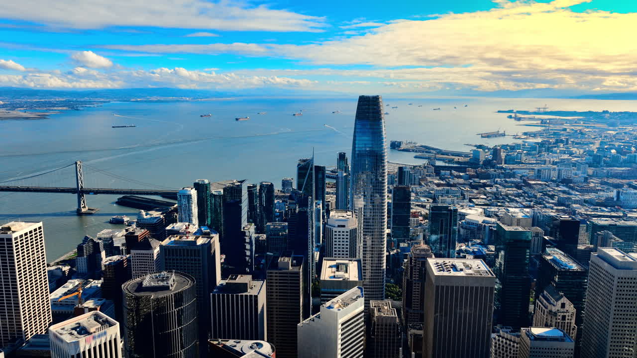 High angle aerial view looking towards the San Francisco Bay and Bay Bridge. The Salesforce Tower surrounded by mixed-use commercial buildings. Ships on the water under a partly cloudy sky