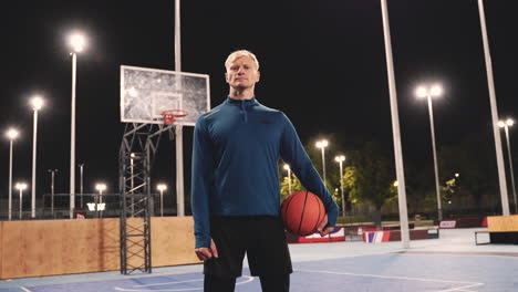 confident basketball player holding ball and looking at camera while standing on an outdoor court at night