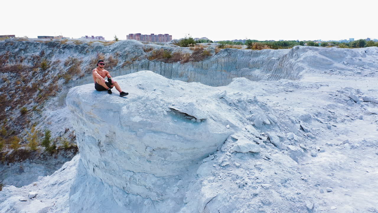 Brutal strong bodybuilder posing outdoor. Photoshoot in a quarry. Outdoor sports concept. Aerial view. Handsome strongman sits on a peak of a quarry. White landscape.
