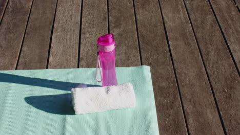 water bottle, towel and yoga mat on sunny wooden deck outdoors, copy space, slow motion