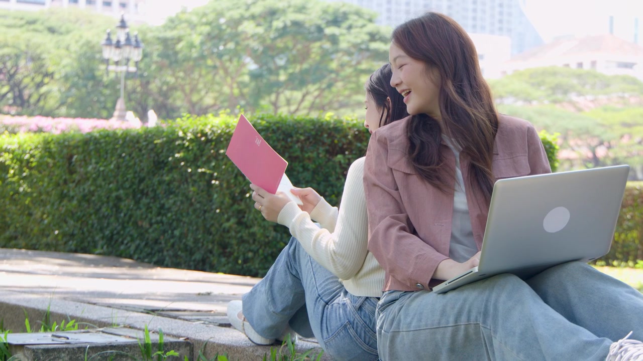 นักเรียนหญิงสองคนนั่งหันหลังชนกันกลางแจ้ง ใช้แล็ปท็อปและสมุดบันทึกในเวลากลางวัน