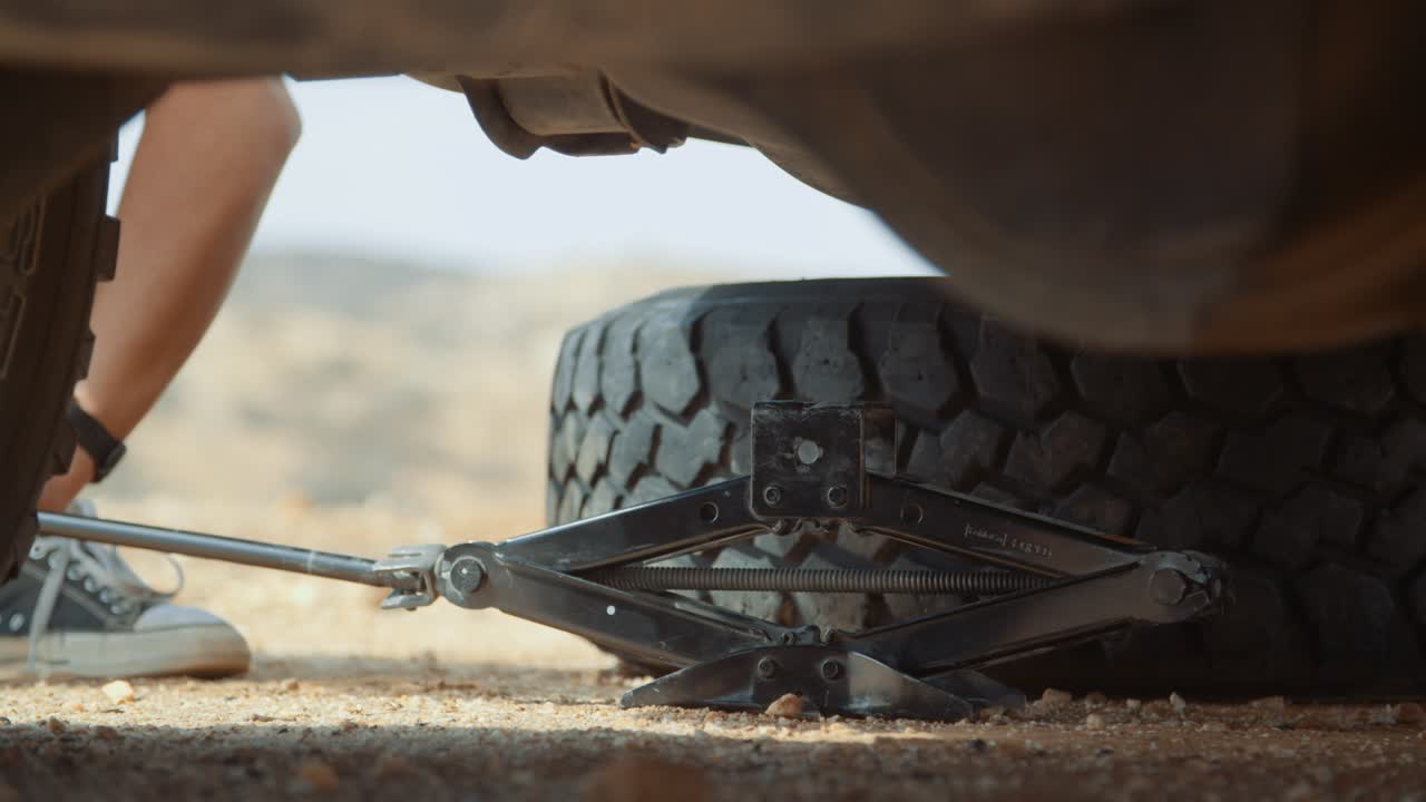 Low angle close-up static  hand held shot of a Caucasian male tourist in Africa as he places a jack underneath a off-roading vehicle with a flat tire