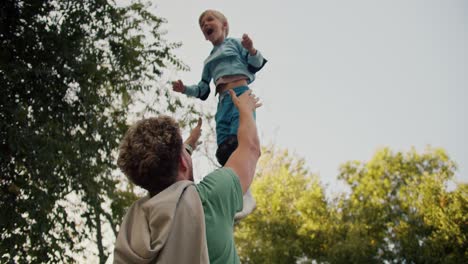 A-father-with-curly-hair-in-a-Green-T-shirt-throws-his-little-blond-son-in-a-Blue-Jacket-up-in-the-park.-A-fun-attraction-for-a-little-boy.-Happiness-in-the-family-between-father-and-son