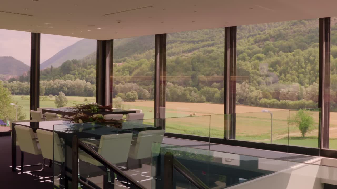 A wide shot pans left, showcasing a modern dining area with white chairs, floral centerpieces, and large windows framing a stunning view of rolling hills and mountains.