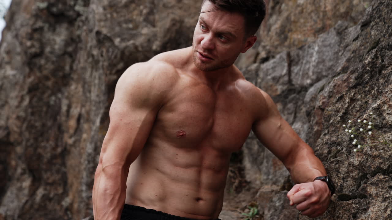 Handsome muscular Caucasian man stands at the rock. Sportsman with naked torso working out in the nature.