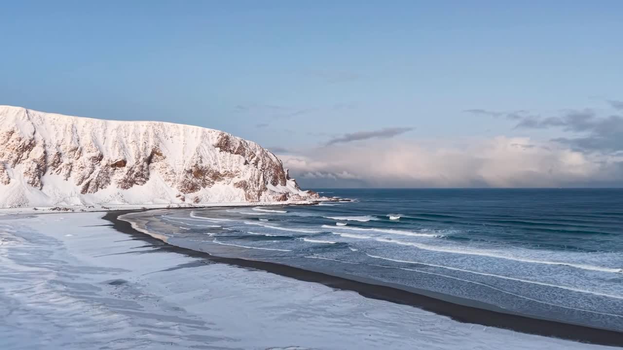 Aerial drone footage of a winter snow covered beach in Norway during a sunny day with a mountain in the background. Ocean sea water is cold blue and has waves with white water, foam and bubbles on it.