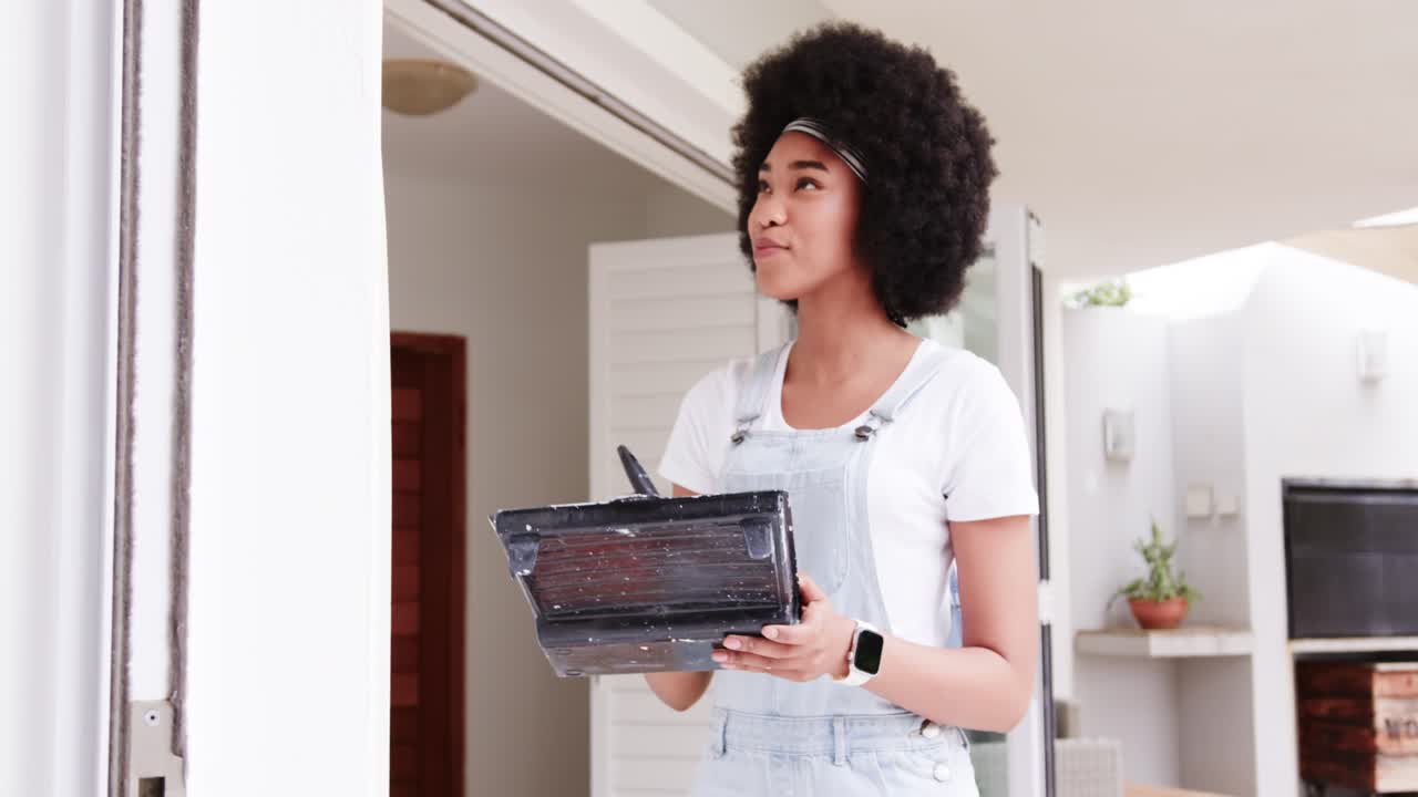 African American woman on porch in overalls surveying above door touching up frame with brush, tray