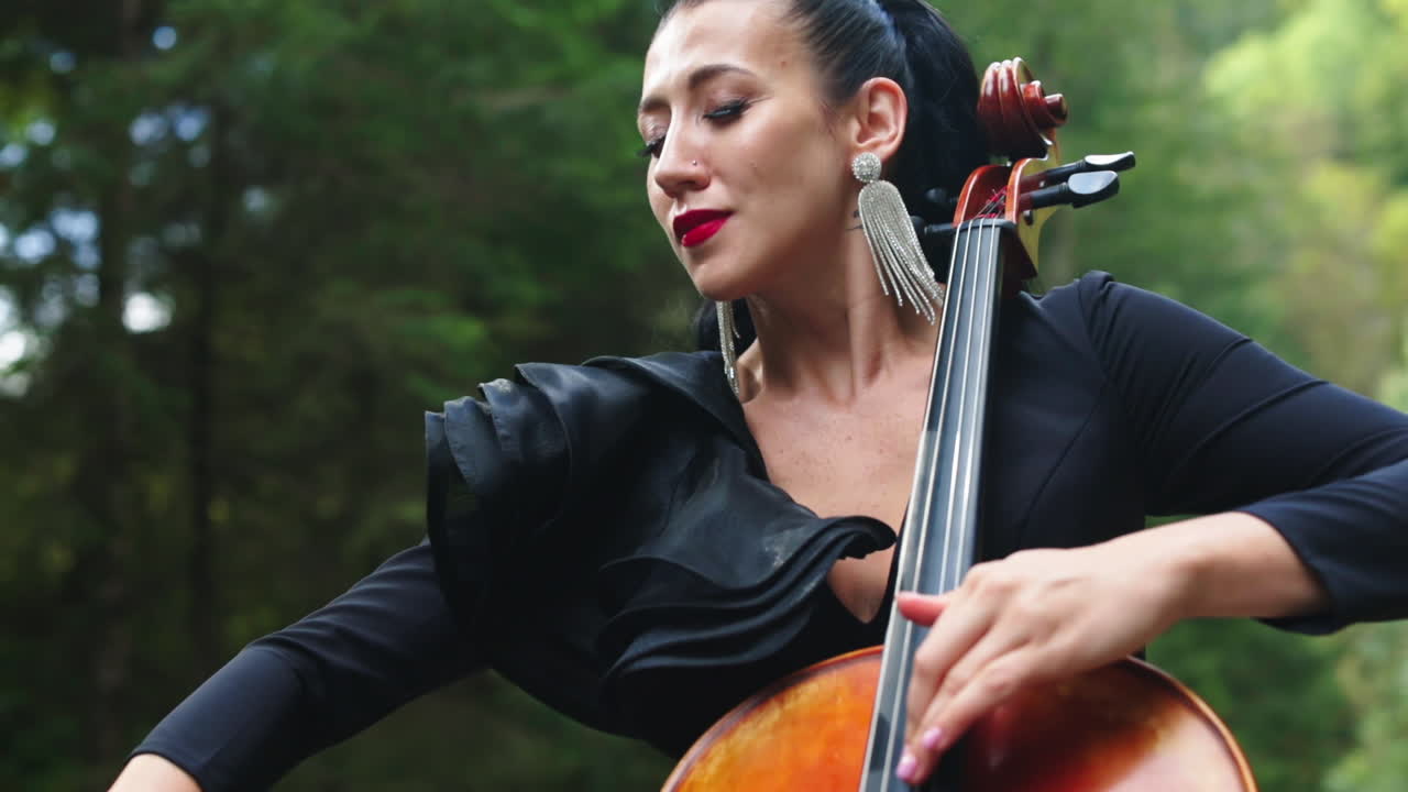 Attractive female musician on green nature background. Gorgeous woman in luxury earrings and dress playing the cello in the green forest.