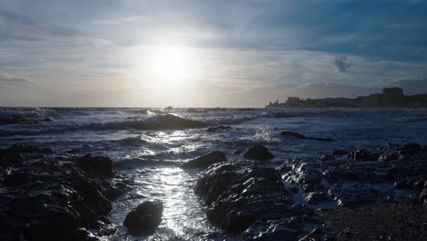 sun reflecting the waves that are hitting the rocks at the beach, with the town of calahonda on the background