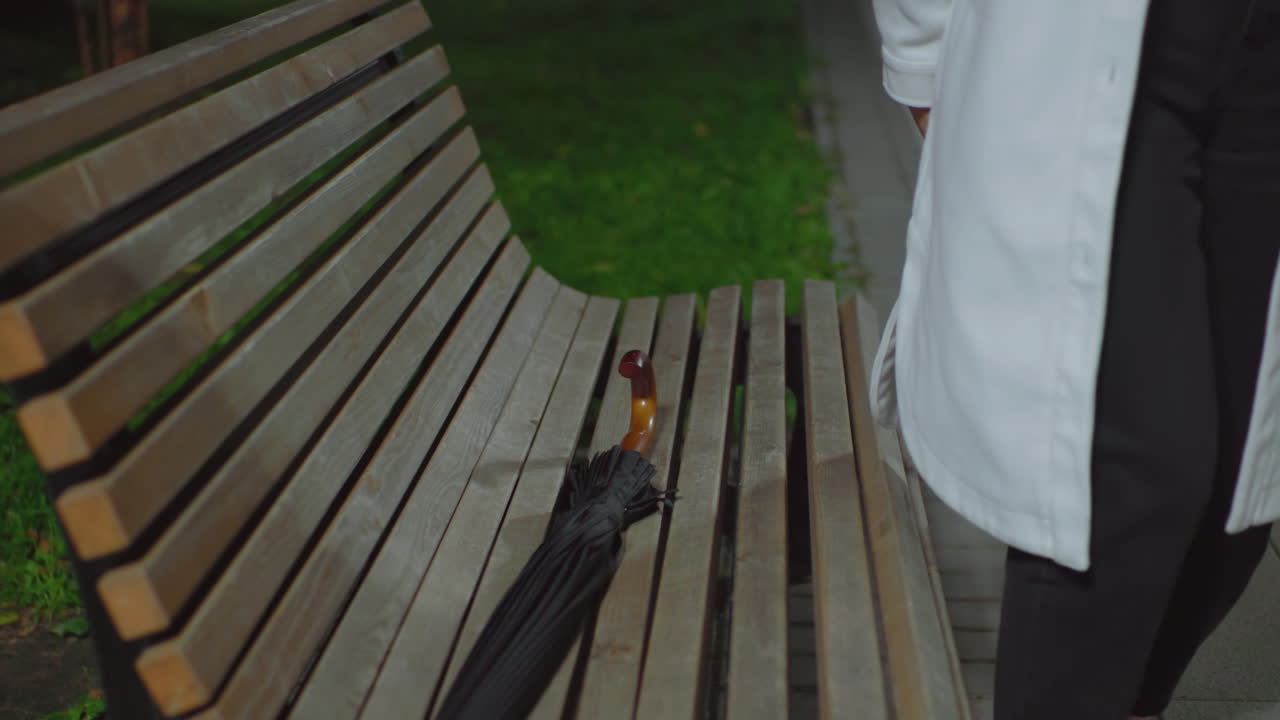 Lady dropping umbrella on wooden bench holding book, preparing to sit down and read during calm night in outdoor park setting, surrounded by greenery and illuminated by soft streetlight glow
