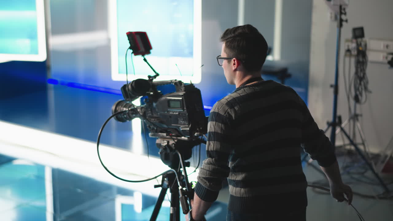 Camera operator adjusting professional video camera setup in modern studio. Focused on filming with studio lights and high-tech equipment, cables, and broadcast production tools visible