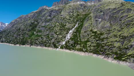 Aerial-drone-tilt-up-shot-of-a-waterfall-along-an-reservoir-in-Grimselsee-along-beautiful-Swiss-Alps,-Switzerland-on-a-bright-sunny-day