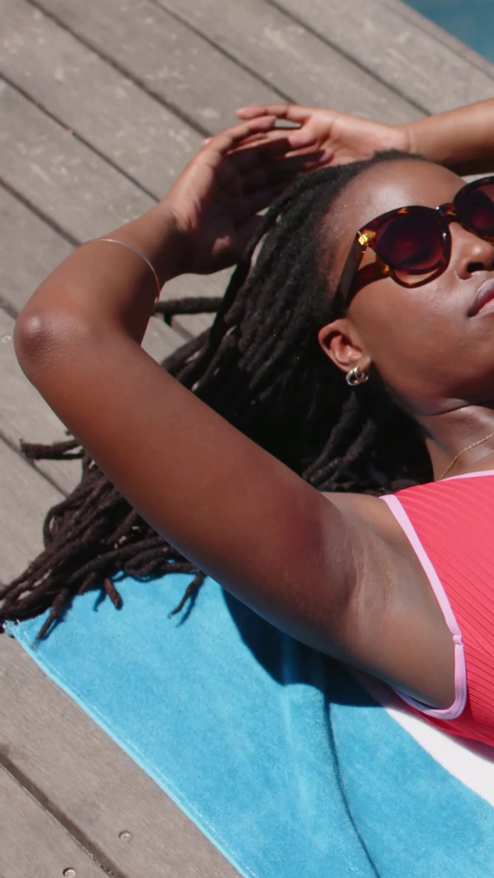 Vertical video: Sunlight nudging woman raising arms on pool towel while relaxing in coral swim top