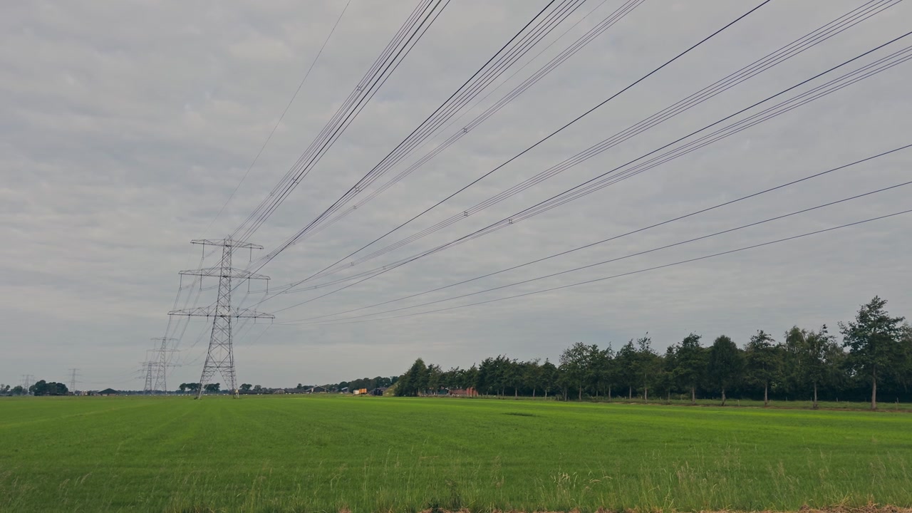 Wide static shot of high-voltage transmission lines and pylons stretching across flat agricultural farmland in the Netherlands. Vibrant green grass contrasts with a moody overcast sky.