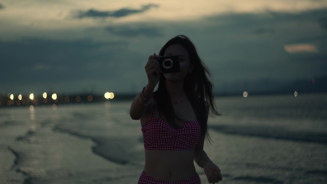 Woman holding a compact camera while standing on the shoreline at twilight, taking photos to capture a peaceful relaxing moment