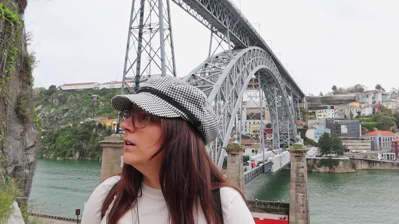 una mujer elegante con gafas y sombrero a cuadros se encuentra frente al ponte luis i, porto