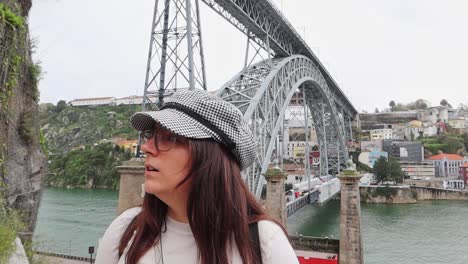 stylish woman in glasses and checkered hat stands in front of ponte luis i, porto