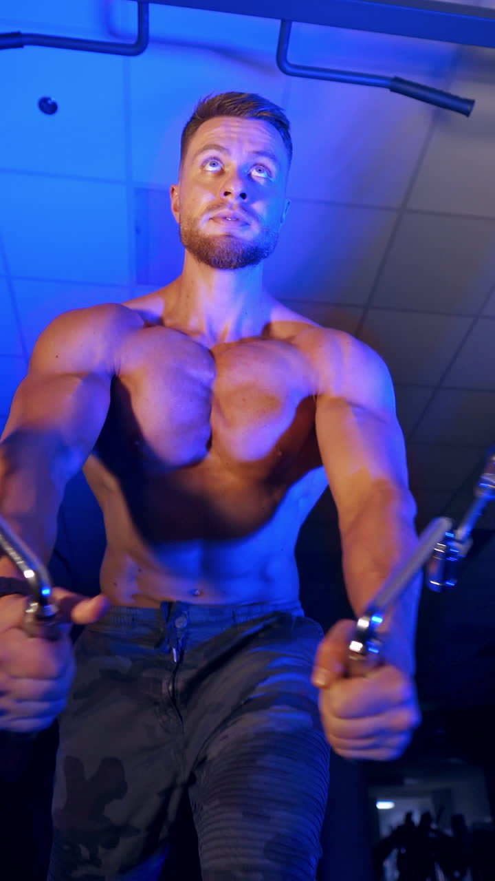 Handsome athlete pumps up his muscles. Naked torso of a sportsman. Muscular bodybuilder training in the gym. View from below. Blue light. Vertical video