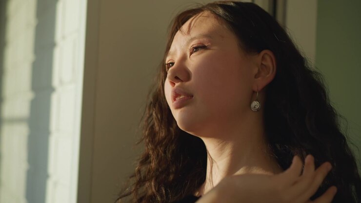 Close up view shows woman with curly dark hair positioned near window illuminated by soft daylight. Her profile is partially visible as woman turns her head slightly revealing texture of her