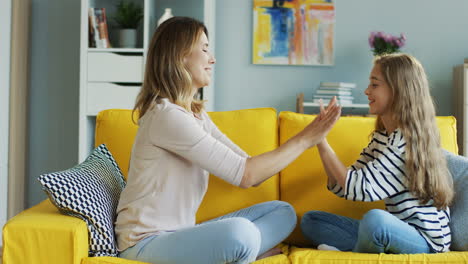 blonde mother with her cute daughter playing hands game while resting on yellow sofa in living room