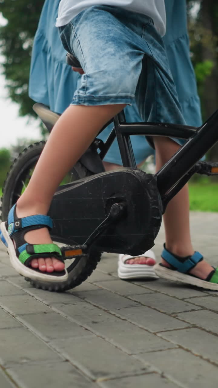 Leg view of a child riding a bicycle, dropping his leg to the ground for balance, while a woman follows closely from behind