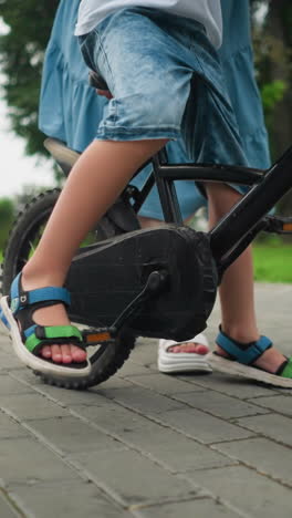 vista de la pierna de un niño montando una bicicleta, dejando caer su pierna en el suelo para el equilibrio, mientras que una mujer sigue de cerca desde atrás