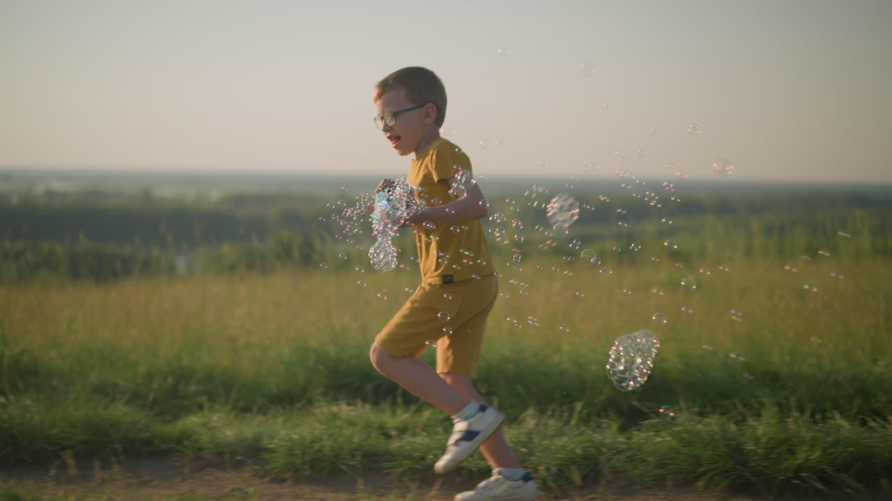 Young boy wearing a yellow shirt and shorts, with glasses, joyfully runs through a sunny grassy field while holding a plastic bubble gun that releases a stream of bubbles