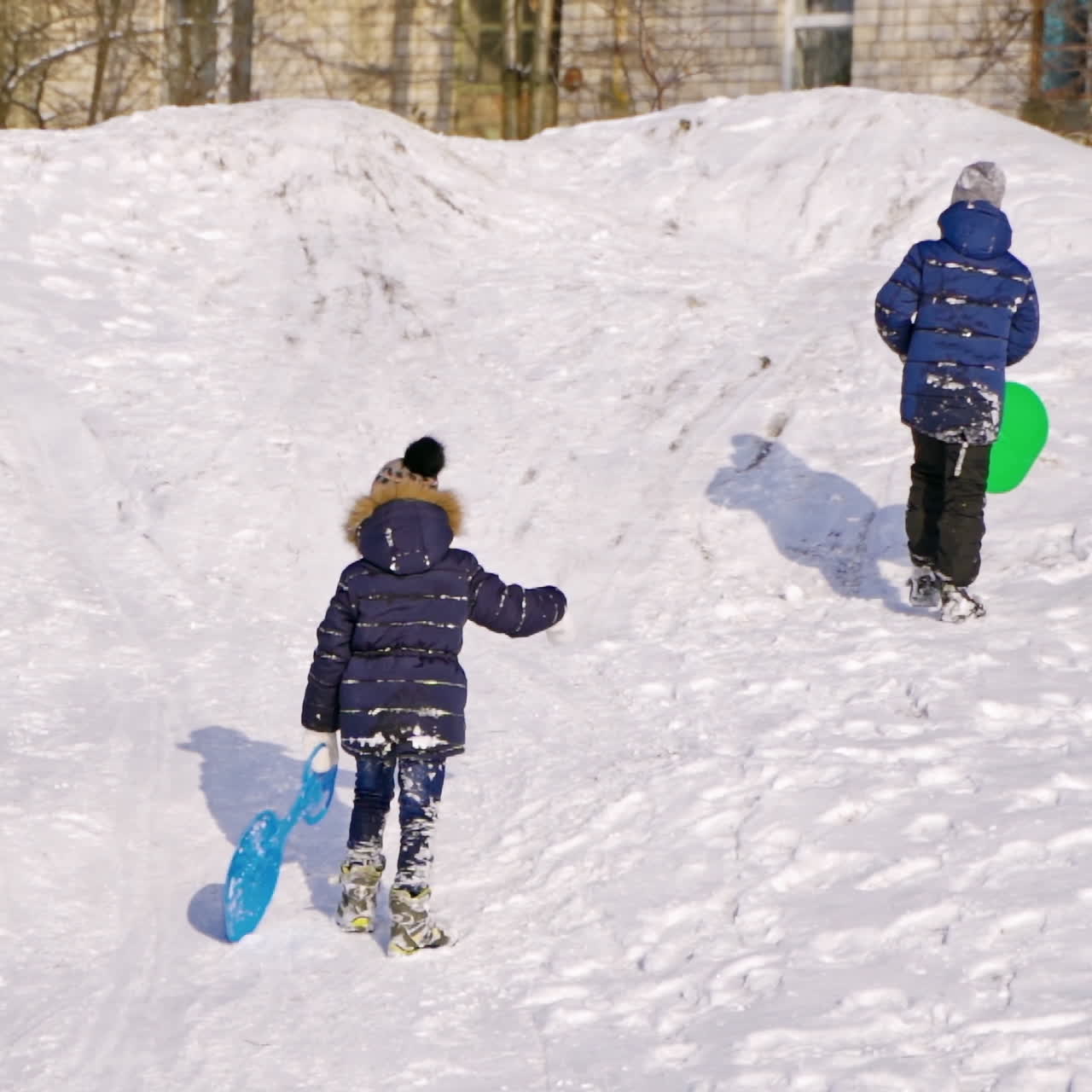 Boys with plastic sleds on a slide in winter. Back view of children on snow background. Slow motion of boys rising up the snowy hill outdoors.