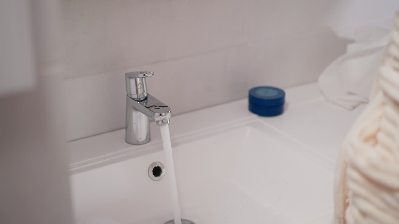 una corriente de agua fluye de la grifa al fregadero en un baño brillante en cámara lenta. la hembra enciende el grifo de agua para lavarse la cara después de la esteticista