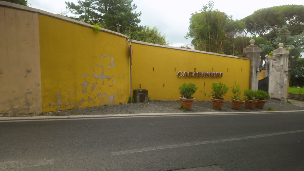 Exterior of a Carabinieri station with yellow wall, potted plants, and tree-lined backdrop in Castel Gandolfo.