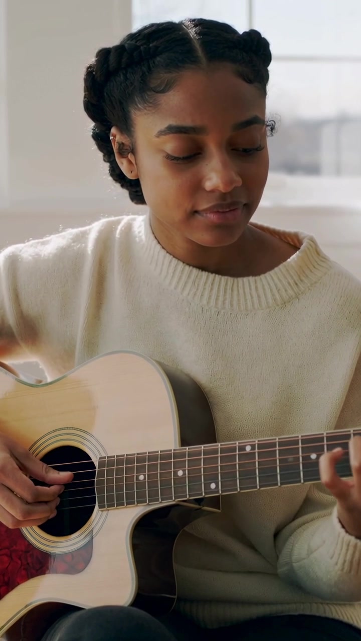 Video verticale: Un musicista seduto posiziona le dita e suona la chitarra a casa, esercitandosi con un maglione color crema.