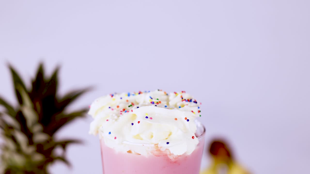 A hand gently places a cherry atop whipped cream and sprinkles on a pink milkshake