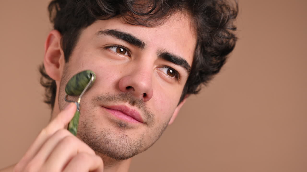 A young caucasian man with stubble beard is doing a facial massage using a roller