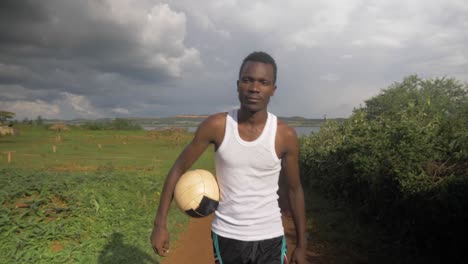 Seguimiento-De-Tomas-En-Cámara-Lenta-Desde-El-Frente-De-Un-Joven-Africano-Caminando-Con-Una-Pelota-De-Fútbol-Bajo-El-Brazo-Por-Las-Tormentosas-Orillas-Del-Lago-Victoria