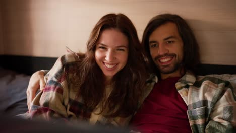 a happy brunette girl in a plaid shirt together with her brunette boyfriend with stubble are watching a movie on a laptop while lying on a bed in a trailer in a camp during their picnic outside the city in the summer