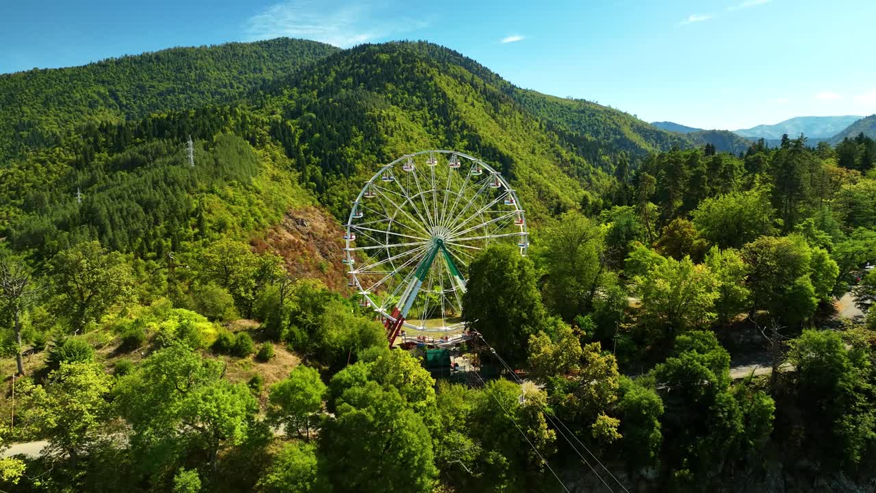 A large ferris wheel rises from a green mountain park, encircled by dense trees and layered hills under bright sunshine