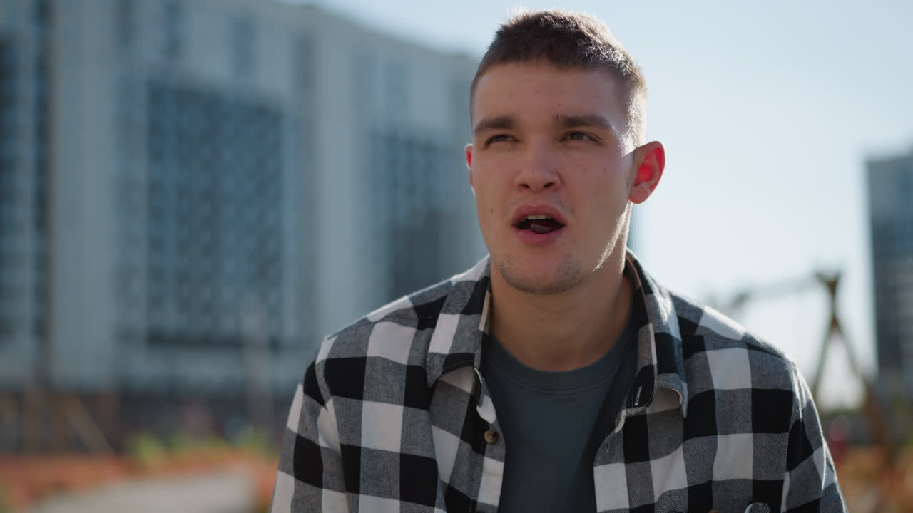 fair skinned man in checkered shirt enjoys sweet while standing outside on bright sunny day with tall residential buildings and wooden playground structures softly blurred in background