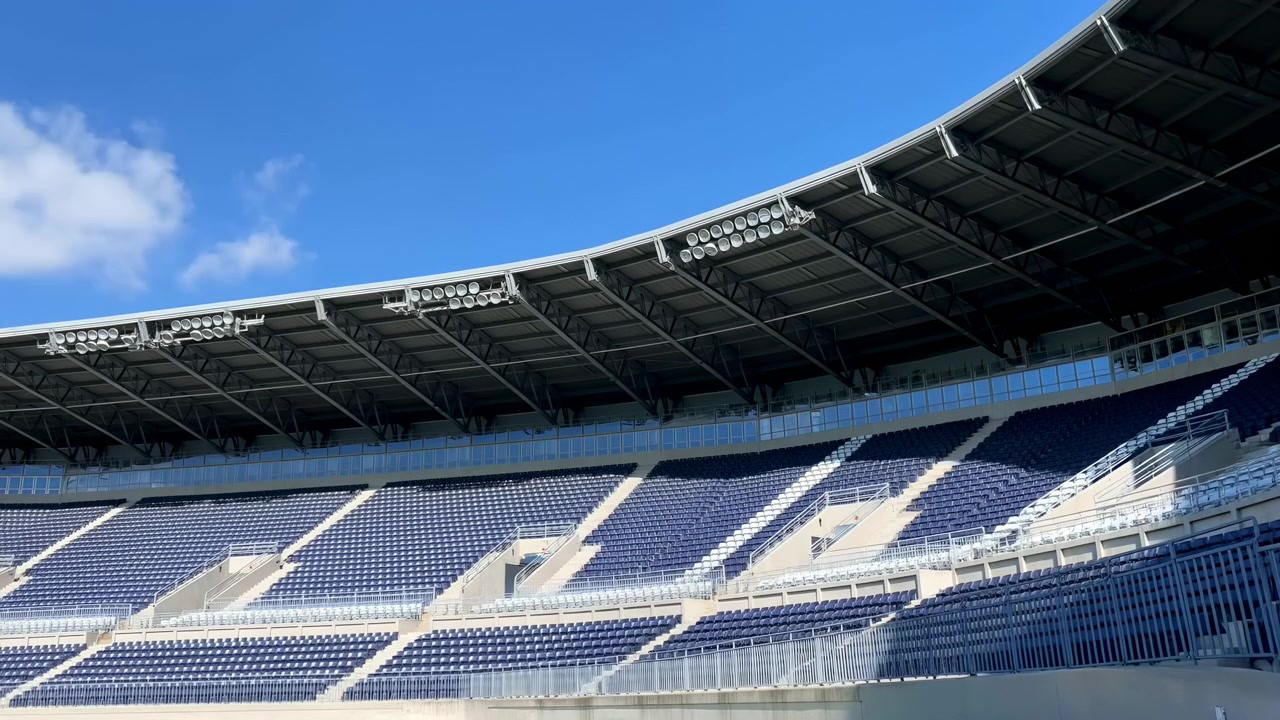 Weitläufige Außenansicht eines großen, leeren Sportstadions mit gestuften blauen Sitzreihen, einem geschwungenen Dach und hellem Tageslicht unter einem klaren Himmel.