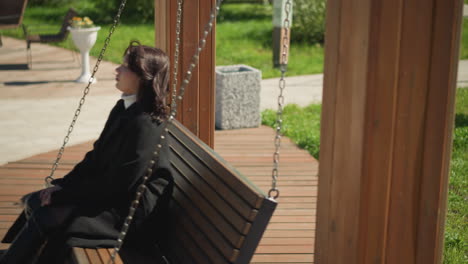 primer plano de una mujer sentada en un columpio de madera, su cabello soplando suavemente en el viento, rodeada de exuberante vegetación en un entorno de parque sereno