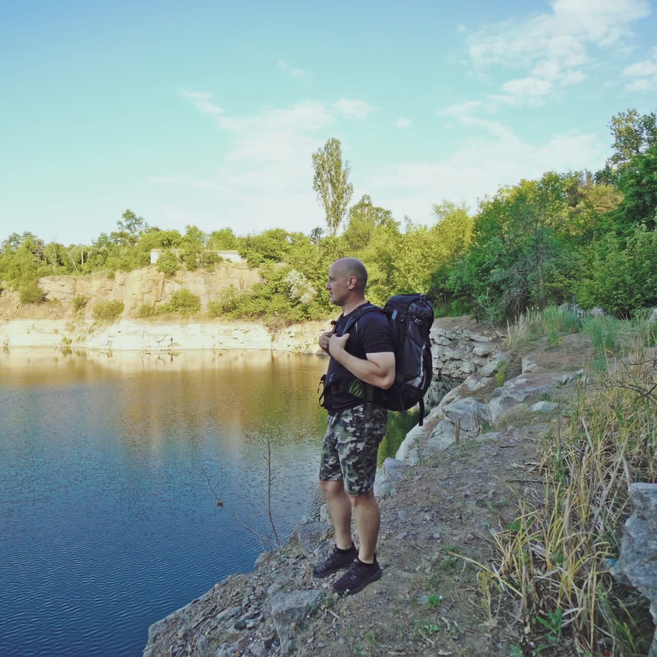 traveler in shorts and a black T-shirt with a backpack on his back is looking at the beautiful view from a cliff by the river in the summer