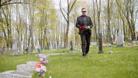 sad man in black suit holding red roses and walking in a graveyard 1