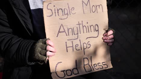 homeless women sitting down holding cardboard sign reading, "single mom, anything helps, god bless"