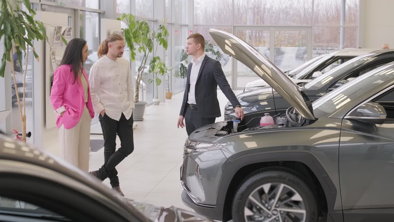 un joven vendedor de coches mostrando a una pareja de jóvenes un nuevo automóvil en el salón de concesionarios.
