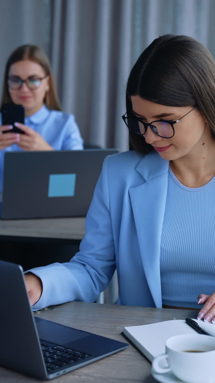 Young female ladies sitting at desks in front of laptops. Employees using gadgets in modern office. Blurred backdrop. Vertical video