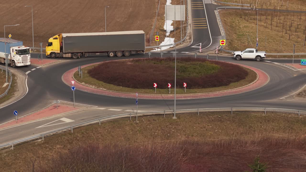 En stor lastbil kör igenom en rondell vid en landsvägskorsning. Flera bilar och skåpbilar navigerar den cirkulära korsningen omgiven av trafikskyltar och vägräcken.