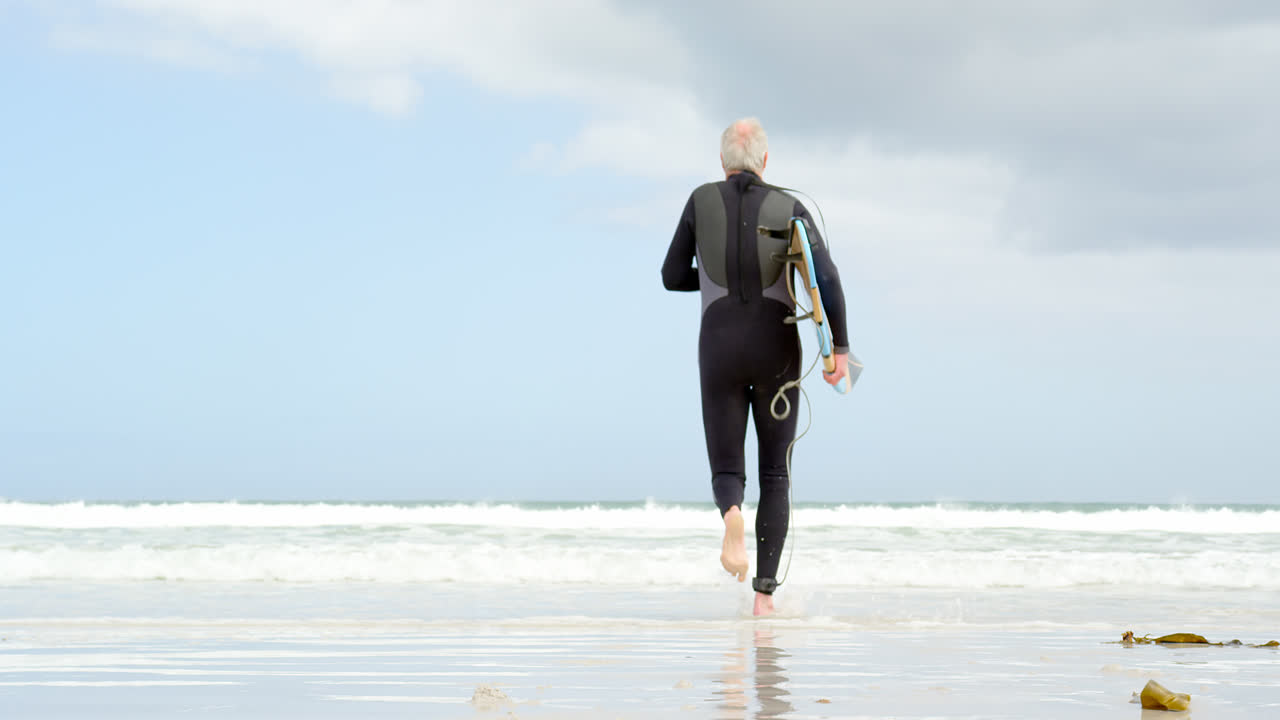 vista trasera de un anciano caucásico corriendo con una tabla de surf en la playa 4k